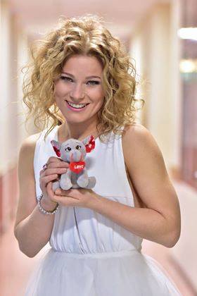 Kobieta w białej sukience trzyma pluszową zabawkę z napisem „Love” i uśmiecha się.