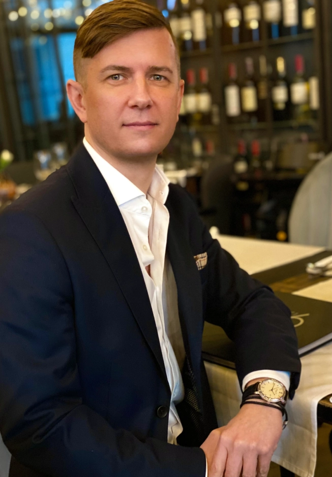 Mężczyzna w garniturze siedzi przy stole w eleganckiej restauracji z winami w tle.