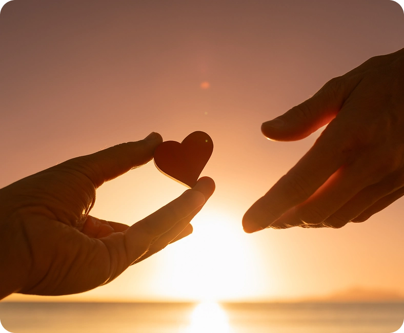 Dwie dłonie przekazują sobie czerwone serce na tle zachodu słońca.
